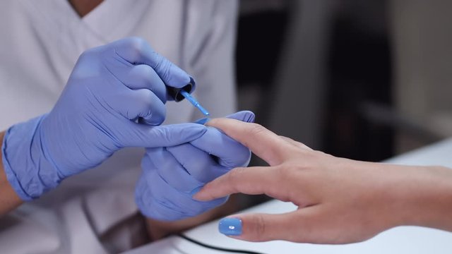 Master wears blue gloves. Nail artist puts blue nail polish using brush. Manicurist makes manicure for female client.