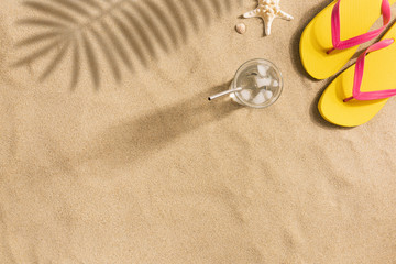 Fototapeta na wymiar Summer fashion, summer outfit on sand background. Yellow flip flops and glass of water. Flat lay, top view. Harsh light with strong shadows