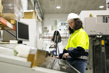 Fototapeta na wymiar Focused female factory worker operating machine at control panel. Middle aged woman in uniform and helmet working at plant. Automated production concept