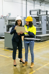 Full length portrait of focused women looking at laptop at plant. Two female employees talking while standing at factory with laptop. Print manufacturing concept
