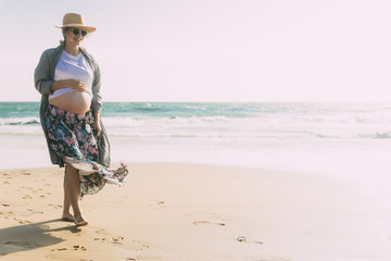 Smiling future mother walking on beach during vacation. Happy pregnant woman holding hand on belly. Pregnancy concept