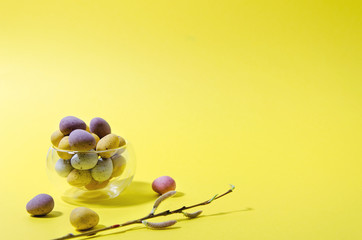 Easter little eggs lie in a glass small mug on a yellow background; next to them are small multi-colored eggs, willow twig and feathers horizontal background minimalism