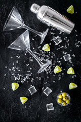 Bar background with martini glasses, shaker, ice, lemon and olives on black table top-down