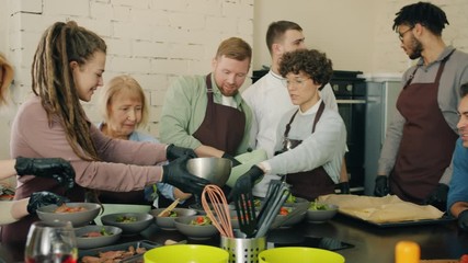 Cheerful cookery class students are working together making meals in modern kitchen wearing aprons and gloves. Food preparation and apprenticeship concept.