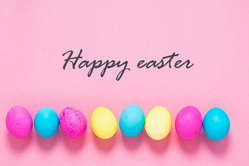 Fototapeta na wymiar Colored eggs lie in a row on pink background text Happy Easter. Festive spring concept. Copy space flat lay banner