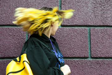 Cute teenager girl with yellow hair and backpack stand behind the brick wall
