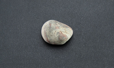 One not big, smooth stone of gray color with brown spots on a black textured background. River pebbles