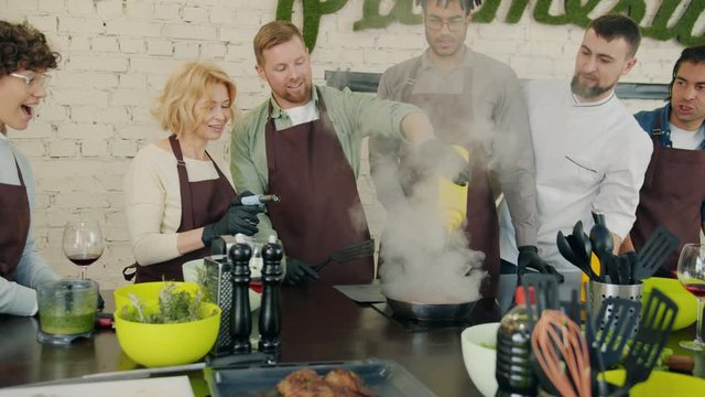 Slow motion of men and women cooking meat with fire in cookery class under guidance of professional male chef, excited trainees are watching flame and smiling
