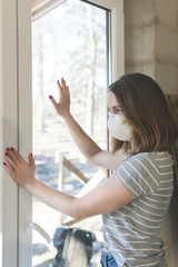 Coronavirus. Young woman staying home in quarantine, looking through the window and wearing protection mask from the illness. Patient isolated to prevent infection of pandemic, epidemic Covid-19 