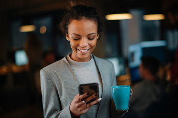 Young businesswoman using phone and drinking coffee in office.	