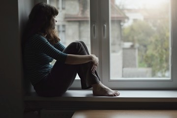 Sad young  beautiful woman sitting on the window at home isolated, watching out. Coronavirus...