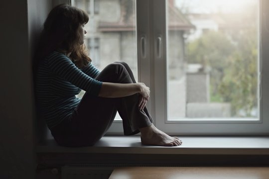 Sad young  beautiful woman sitting on the window at home isolated, watching out. Coronavirus quarantine concept.