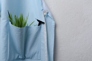Closeup of blue garden apron and pocket of it full of green grass and little shovel, rake, white wall.Empty space