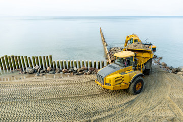 an excavator on the beach loads sand and stones into the truck body, works to strengthen the beach and install breakwaters