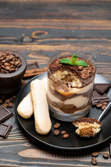 Classic tiramisu dessert in a glass cup, pieces of chocolate and savoiardi cookies on wooden background