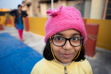 Low angle portrait of an Indian brunette baby girl with woolen clothes, cap and spectacles doing funny facial expression in a casual mood on a rooftop in winter afternoon. Indian lifestyle.