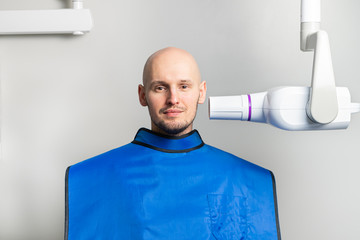 dental x-ray room. Portrait of a young bald man in a protective lead apron next to an X-ray machine during diagnosis.