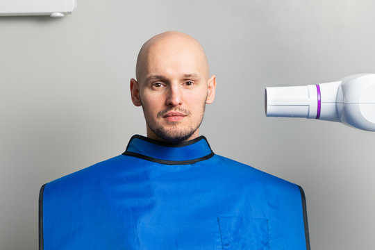 Young Bald Man In A Blue Protective Lead Apron In An X-ray Room Near An X-ray Apparatus For Dental Imaging. Dentistry