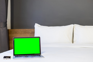 Laptop computer with green screen with smart phone on white bed decoration in hotel bedroom interior background,Work and business in leisure with travel in the holiday concept.