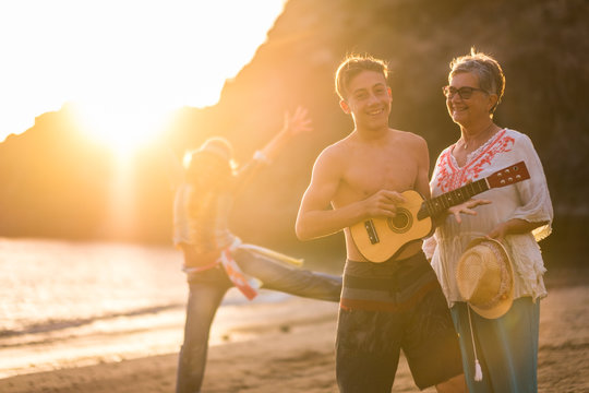 Happy family have fun at the beach with a guitar - mixed generations from grandson to grandmother and mother together having happiness - playful people enjoy summer holiday vacation
