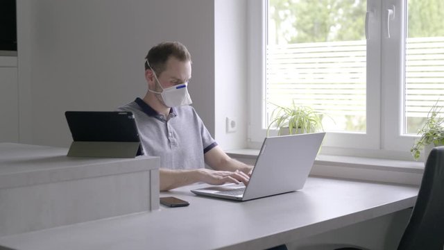 Man wearing sanitary surgical face mask and working on laptop in front of a window while getting a phone call