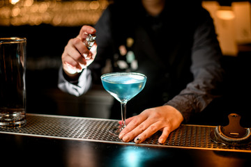 blue alcoholic cocktail in wineglass stands on bar counter. Bartender's hand near glass.