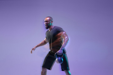 Fototapeta na wymiar Emotions. Young african-american bodybuilder training over purple background in neon, mixed light. Muscular male model with weight. Concept of sport, bodybuilding, healthy lifestyle, motion and action
