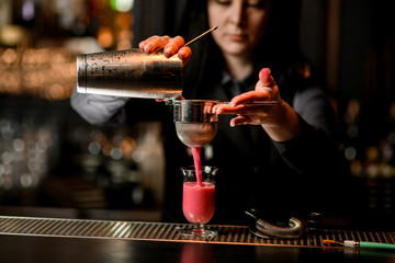 Fototapeta na wymiar brunette barman gently pours cocktail from steel shaker into glass using sieve.
