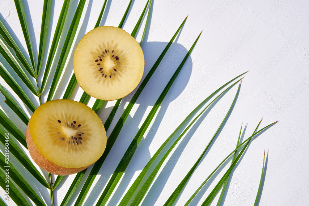 Wall mural Green palm leaf with two halves of ripe golden kiwi laying on it, close-up shot from above on white natural background in harsh sunlight with colored shadows.