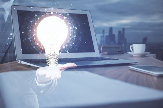 Computer on desktop in office with bulb icon hologram. Multi exposure. Concept of idea.