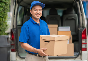 A smiling young asian delivery in blue uniform with parcel cardboard in front of customer house. Messenger and delivery concept.