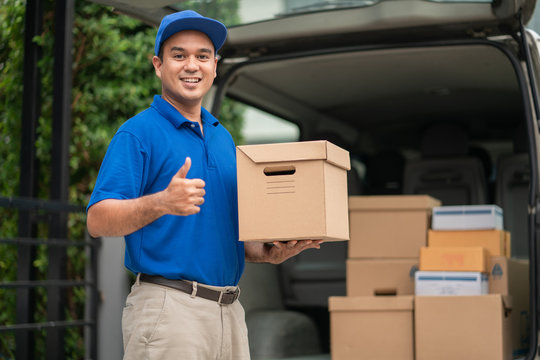 A smiling young asian delivery in blue uniform with parcel cardboard in front of customer house. Messenger and delivery concept.