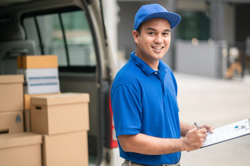 A smiling young asian delivery in blue uniform with parcel cardboard in front of customer house. Messenger and delivery concept.