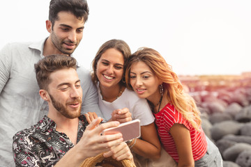 Young friends sitting outdoors and looking at smartphone. Young man showing something interesting to friends outdoors at the seaside