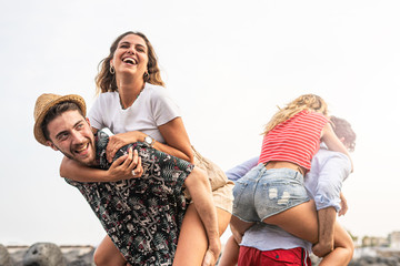 Group of friends walking along the seaside, with men giving piggyback ride to girlfriends. Happy...