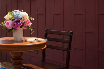 Artificial rose flowers in white ceramic vase, pen, notebook on wooden round table with teak wood chair and wooden wall background in vintage style, side view with copy space