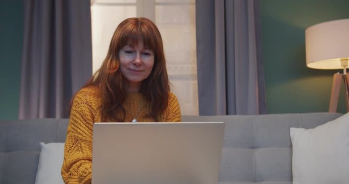 Smiled Caucasian red-haired woman sitting on sofa in living room with lapop computer while working on it or surfing online indoor. Beautiful female homey typing on keyboard.