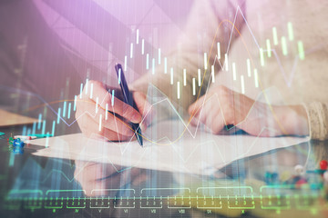 Hand taking notes in notepad. Forex chart holograms in front. Concept of research. Multi exposure