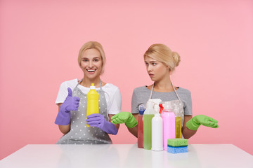 Indoor shot of young white-headed cleaning ladies sitting over pink background in working clothes, one getting best detergent and another has a lot of worse bottles