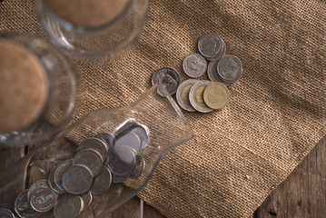 Thai Baht coins poured from glass bottle on gunny sack on wooden table, top view, saving and emergency fund concept