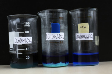 Three beakers with complex inorganic compounds: dark tetraammine cobalt sulfate, blue precipitate tetraammine cuprosulfate, blue liquid tetraammine nickelsulfate, with formulas, on a black background.