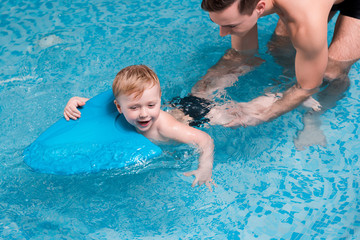 excited toddler boy swimming with flutter board near swim coach