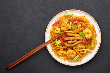 Singapore Mei Fun in white plate on dark slate background. Singapore Noodles is chinese cuisine dish with rice noodles, prawns, char siu pork, carrot, red onion, napa cabbage. Chinese food. Copy space