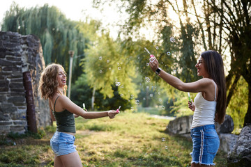 Naklejka na ściany i meble Two sisters, blowing soap bubbles at each other in park, laughing, smiling, Young pretty girls, wearing jeans shorts, green beige tops, having fun outside. Three-quarter portrait of two girlfriends.