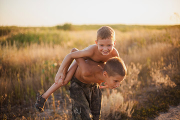 The older brother carries his laughing younger brother on his back in the summer field at sunset.