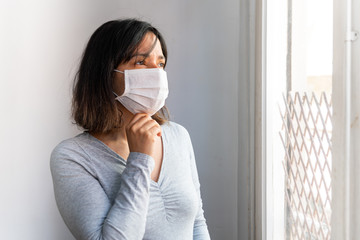portrait of young woman with face mask in quarantine due to corona virus inside her home with sad...