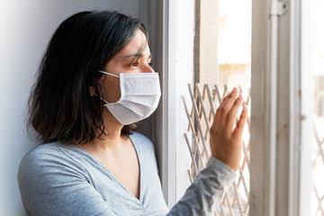 portrait of young woman infected with corona virus with face mask looking out of window while in quarantine with sad and worried expression due to isolation - obrazy, fototapety, plakaty