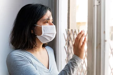 Fotobehang portrait of young woman infected with corona virus with face mask looking out of window while in quarantine with sad and worried expression due to isolation © davide bonaldo