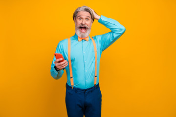 Photo of cool shocked grandpa hold telephone open mouth surprised new model smart phone wear blue shirt suspenders bow tie pants isolated yellow color background