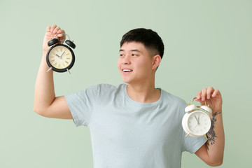 Young Asian man with alarm clocks on color background